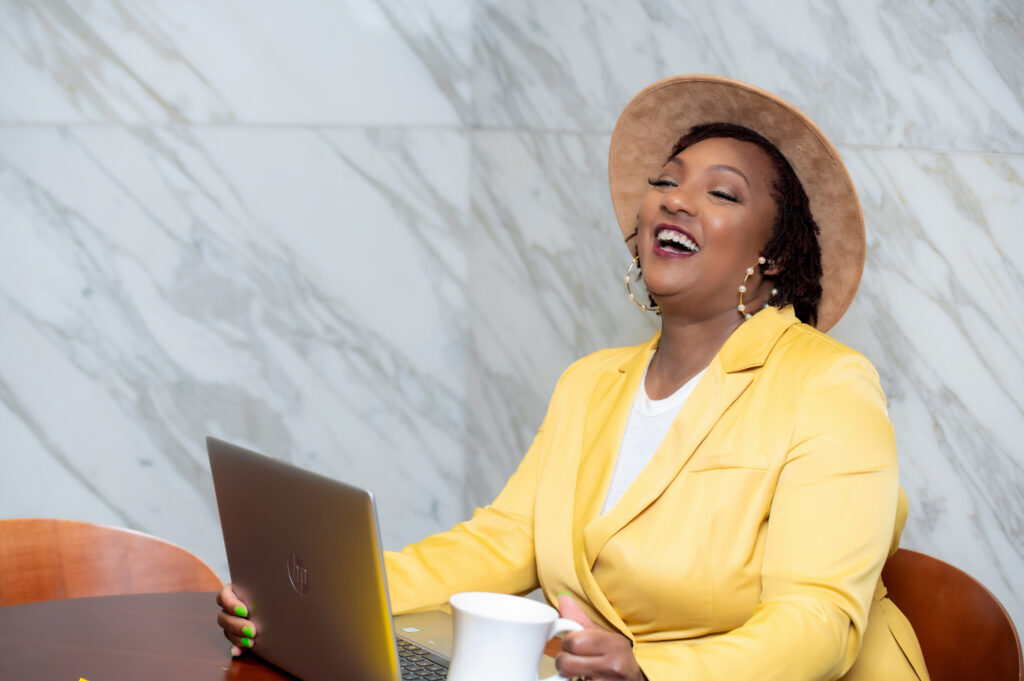 Smiling black woman sitting at desk in yellow blazer and fedora using a computer and holding a white coffee mug